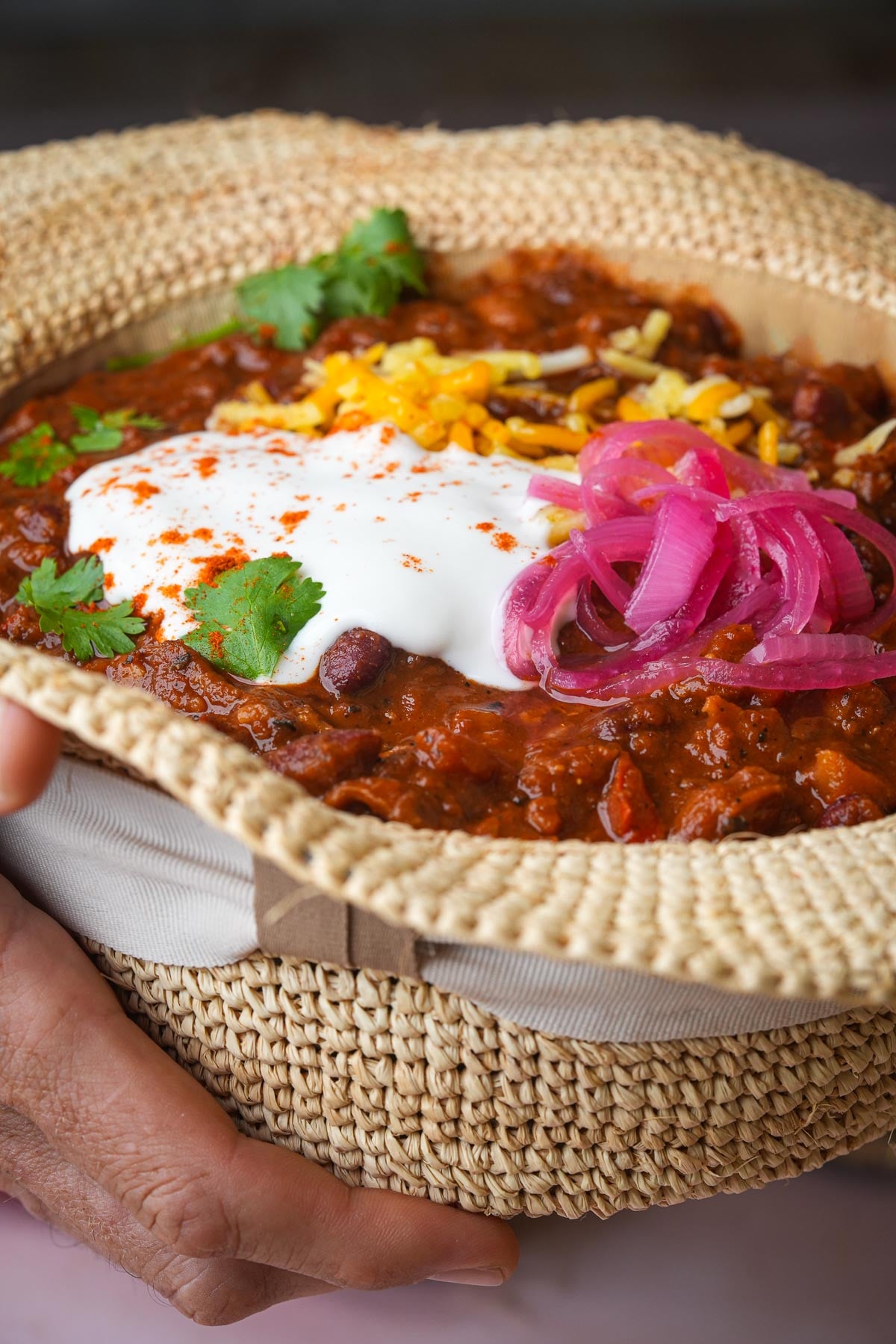 Person holding a woven hat filled with vegan chili topped with shredded vegan cheese, sour cream, sliced red onions, and cilantro.