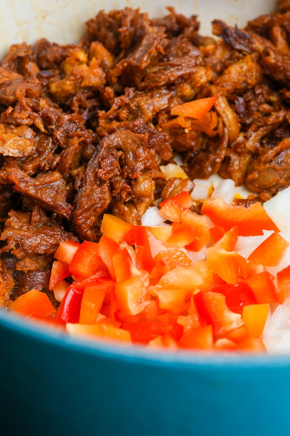A close-up of shredded seitan and diced red bell peppers in a blue cooking pot.