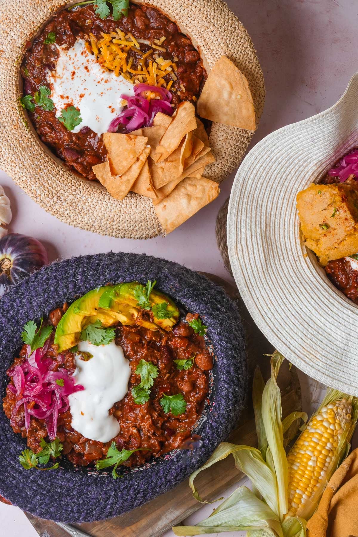Three bowls of chili garnished with sour cream, avocado slices, shredded cheese, red onions, and tortilla chips, placed in sun hats. Corn cobs, garlic, and herbs are visible around the bowls.
