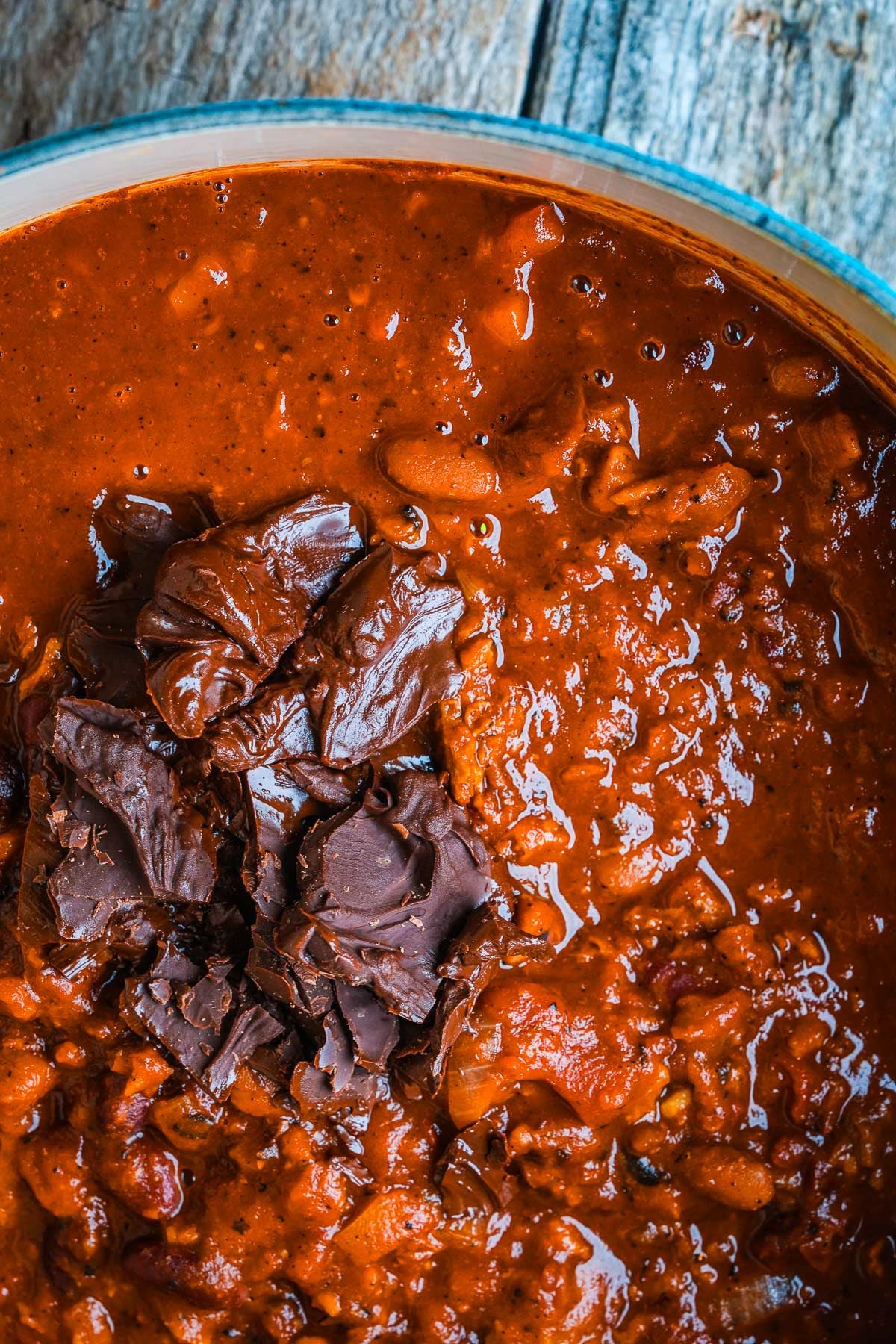 A close-up of a bowl of vegan chili topped with pieces of dark chocolate. The background is wooden.