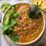 A bowl of chunky salsa topped with a charred green pepper and fresh cilantro, with hints of salsa verde and tortilla chips visible in the background.