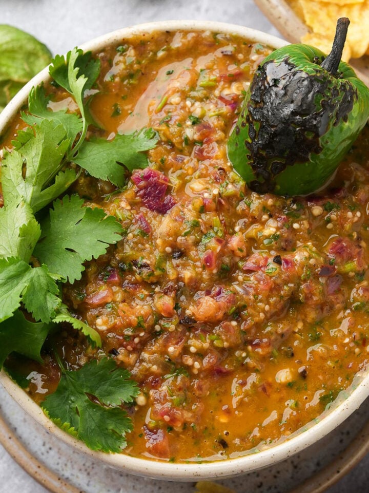 A bowl of chunky salsa topped with a charred green pepper and fresh cilantro, with hints of salsa verde and tortilla chips visible in the background.