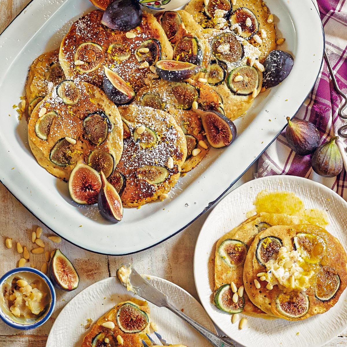 A platter and two plates of pancakes topped with figs, pine nuts, and powdered sugar are displayed on a table with a cloth and whole figs. A small bowl with additional toppings is also present.