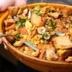 A close-up of a bowl of vegan stuffing with bread cubes, celery, herbs, nuts, and slices of mushrooms, being held by two hands.
