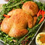 A roasted baby-shaped tofu loaf on a baking tray, surrounded by green leafy vegetables, carrots, and sprigs of herbs. A small bowl of dipping sauce is placed nearby.