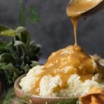 A ladle pours Vegan Brown Gravy over a serving of mashed potatoes in a bowl, with herbs in the background.
