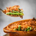 A spoon lifting a serving of green bean casserole topped with crispy fried onions from a brown baking dish.