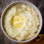 A bowl of vegan mashed potatoes topped with melted butter, black pepper, and chopped chives on a wooden surface.