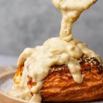 A flaky pastry topped with sesame seeds is being covered with a thick, creamy vegan sausage gravy.