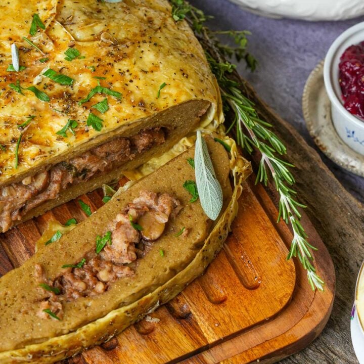 A sliced vegan turkey roast, garnished with fresh herbs, on a wooden board. Several sprigs of rosemary and a small bowl of sauce are visible in the background.