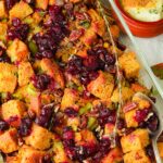 A baking dish filled with vegan cornbread stuffing, featuring chunks of cornbread, cranberries, pecans, and celery. A metal serving spoon rests on the dish. A small bowl with dipping sauce is nearby.