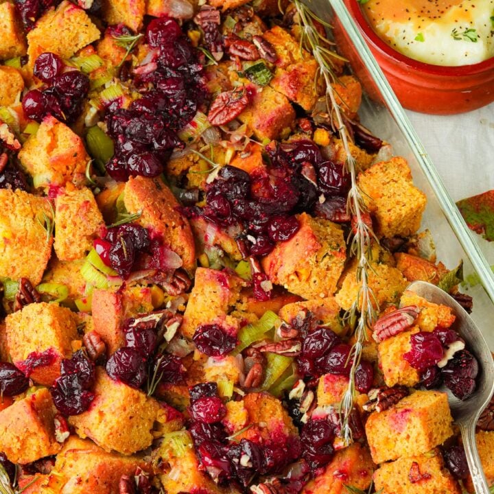 A baking dish filled with vegan cornbread stuffing, featuring chunks of cornbread, cranberries, pecans, and celery. A metal serving spoon rests on the dish. A small bowl with dipping sauce is nearby.