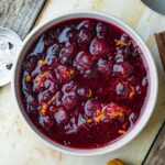 A white bowl filled with dark red bourbon cranberry sauce with chunks of orange zest mixed in. The bowl is placed on a rustic wooden surface.