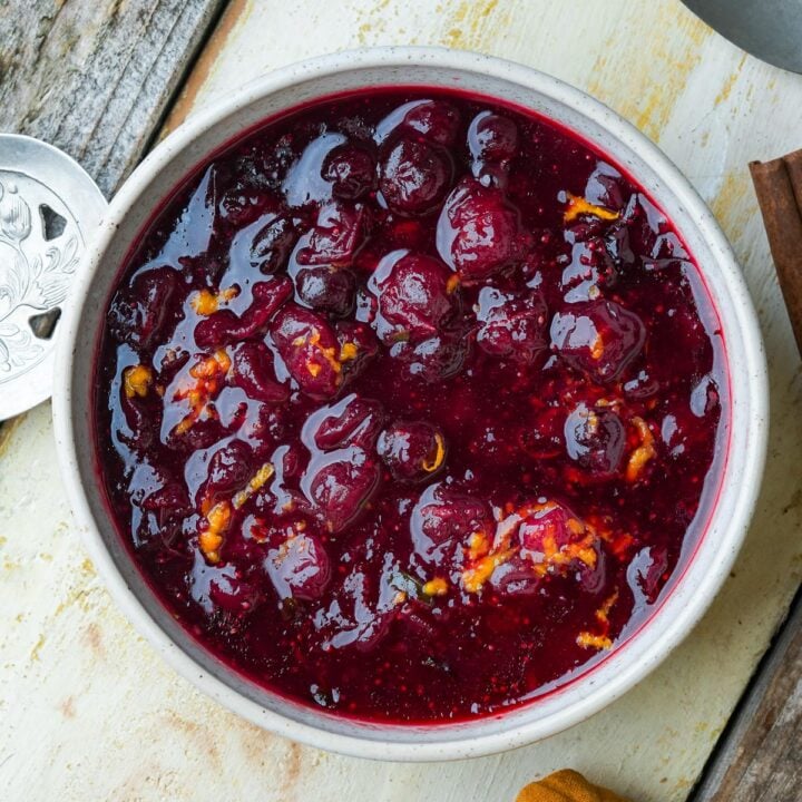 A white bowl filled with dark red bourbon cranberry sauce with chunks of orange zest mixed in. The bowl is placed on a rustic wooden surface.