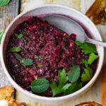 A bowl of red Cranberry Ezme garnished with fresh mint and parsley, with a spoon on the side. Toasted bread pieces are visible nearby.