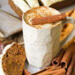 A textured mug filled with frothy vegan eggnog, topped with a cinnamon stick. Slices of bread and more cinnamon sticks are on the side.