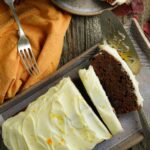 A sliced vegan gingerbread loaf cake with white frosting on a ceramic plate, accompanied by a cake server and a fork on an orange napkin.