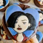 A hand holding a chocolate-covered treat with a cartoon character's face, featuring black hair and a blue background, depicted on it.