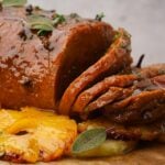 Close-up of a glazed roast vegan ham garnished with fresh herbs, accompanied by caramelized pineapple slices on a wooden board.