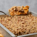 A serving of vegan sweet potato casserole with a crumbly topping is being lifted from a baking dish.