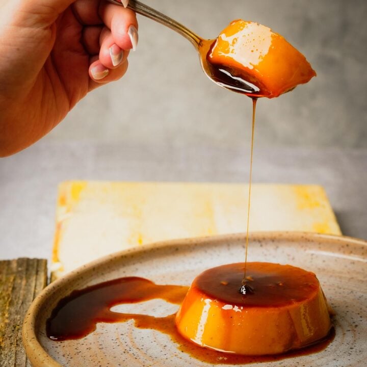 A hand holds a spoon with a piece of Pumpkin flan, allowing caramel sauce to drip onto the dessert on a plate below.