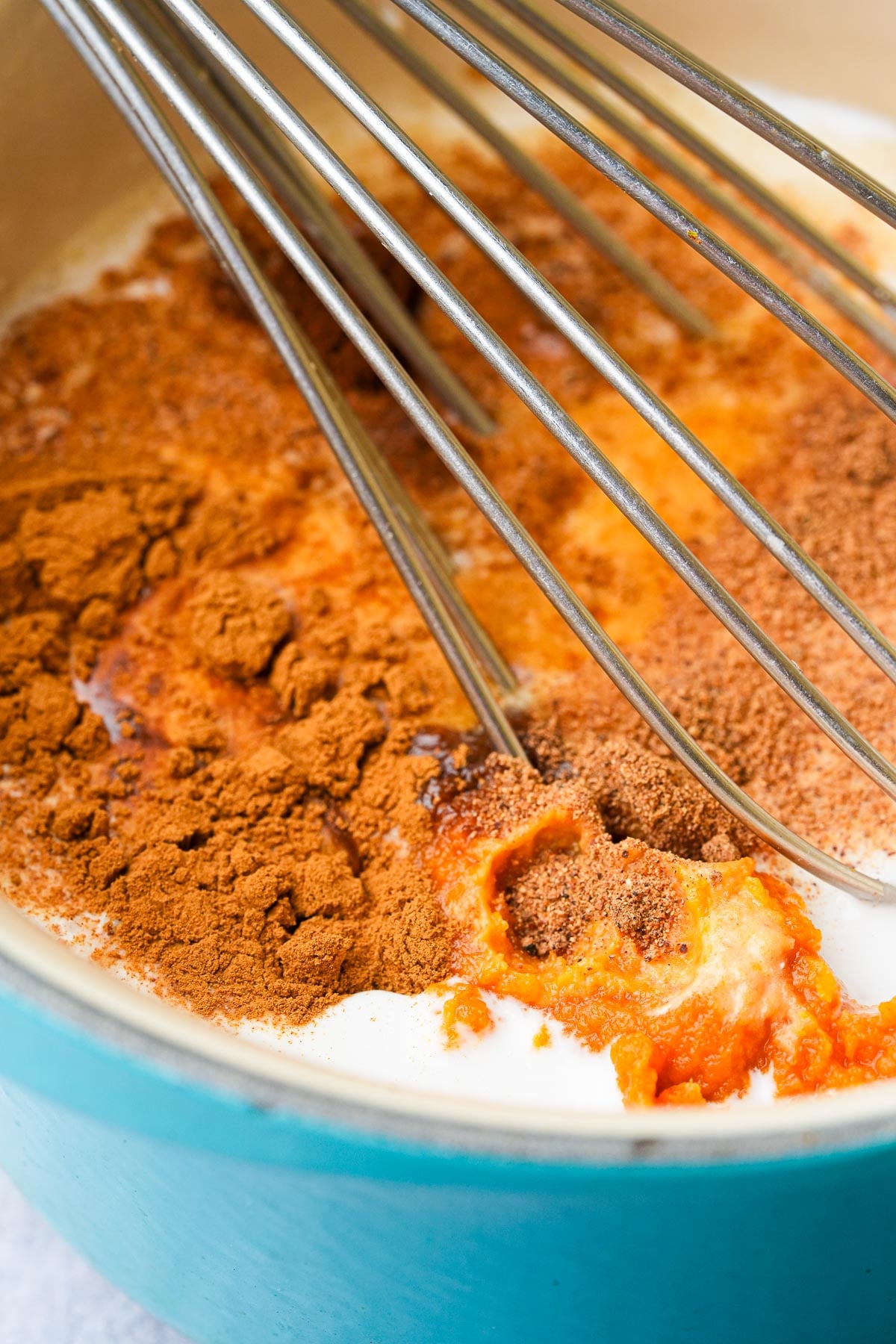 Close-up of whisks mixing pumpkin puree, milk, cinnamon, and other spices in a blue saucepan.
