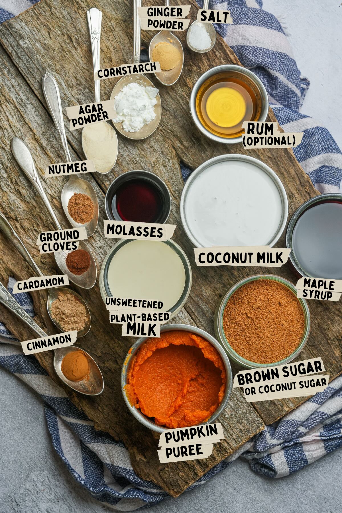 A wooden surface with various Pumpkin flan ingredients labeled: ginger powder, salt, cornstarch, agar powder, nutmeg, rum, molasses, coconut milk, plant-based milk, maple syrup, pumpkin puree, spices, and sugar.