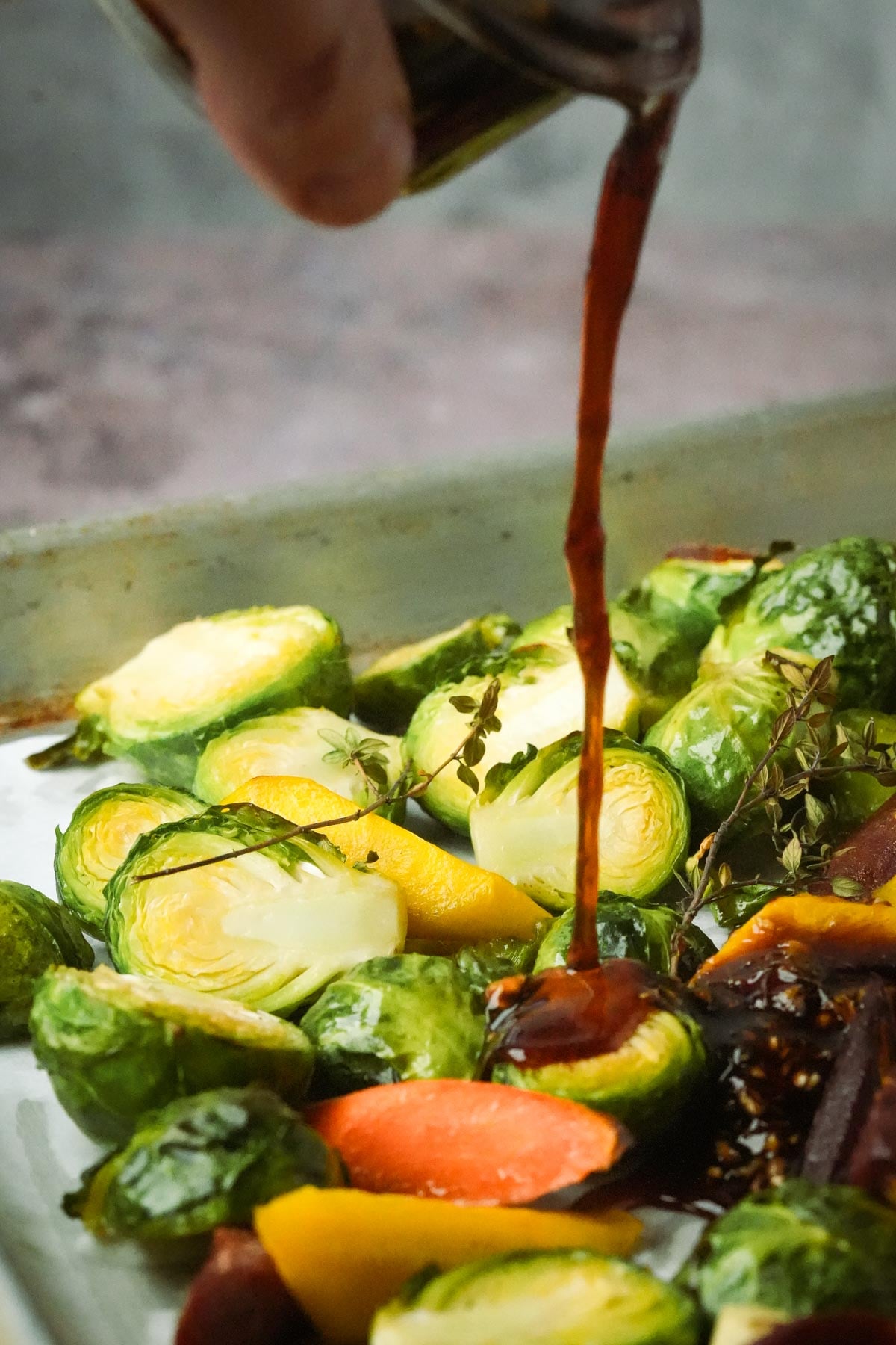 Pouring balsamic maple glaze over roasted Brussels sprouts, carrots, and herbs on a baking sheet.