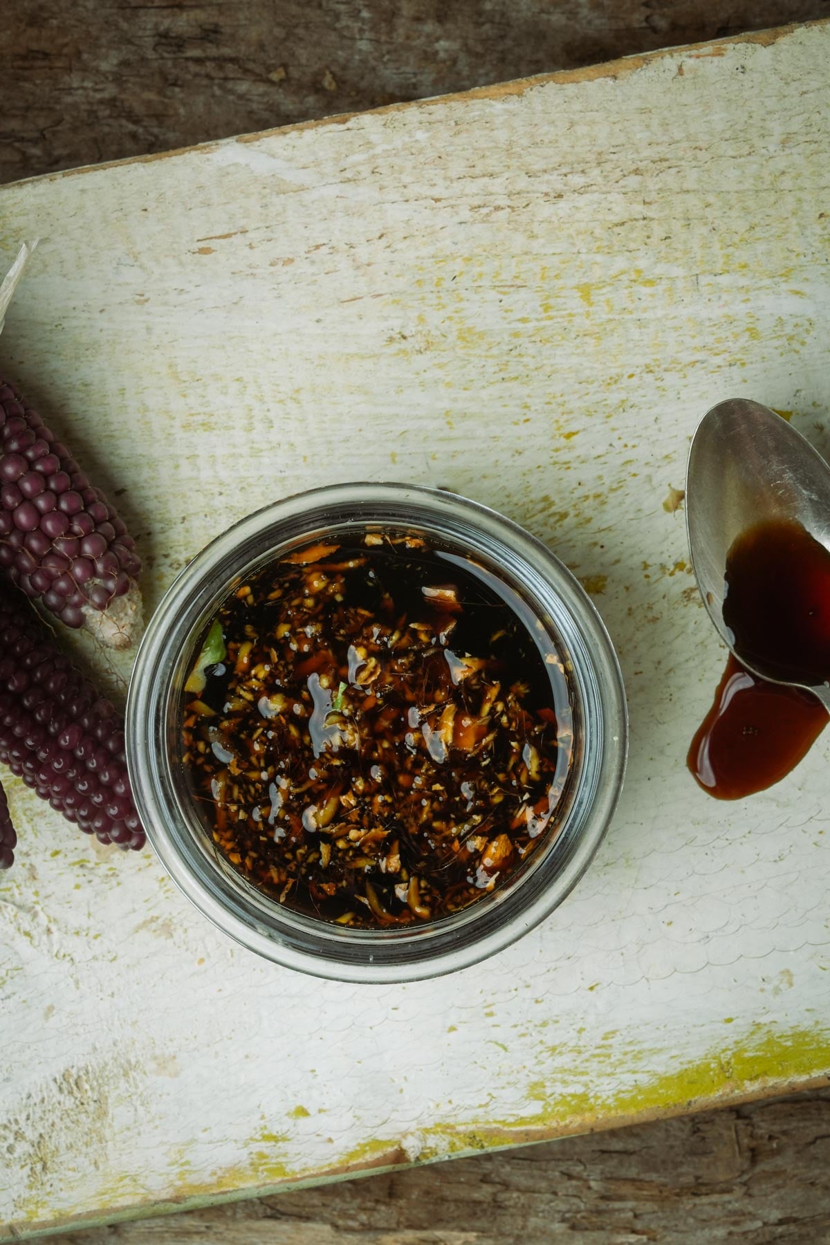 Top view of a glass jar filled with maple balsamic glaze with ginger, next to two purple corn cobs and a spoon with sauce dripped on a white surface.
