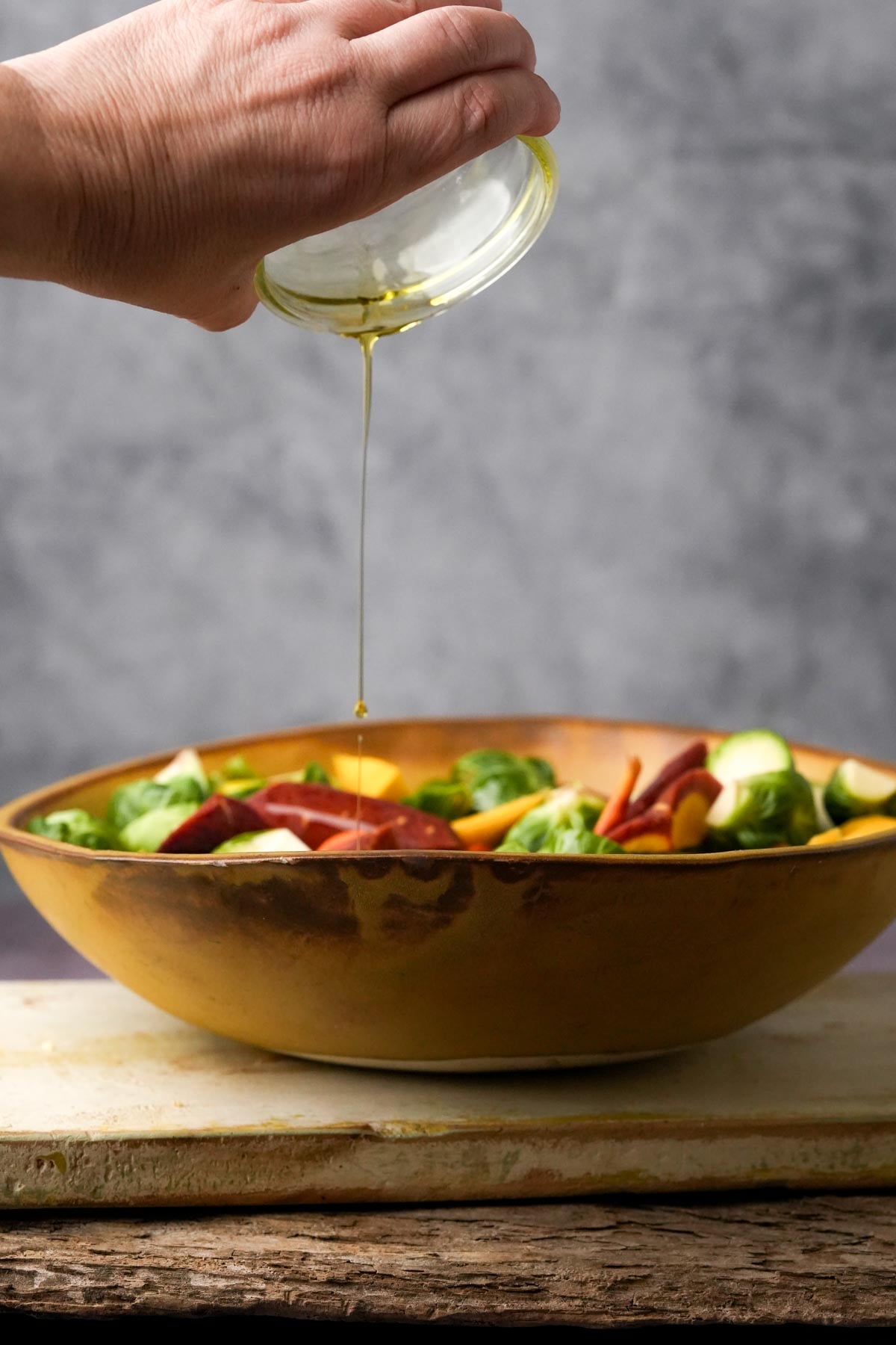 A hand pouring olive oil into a ceramic bowl of mixed vegetables on a wooden surface.