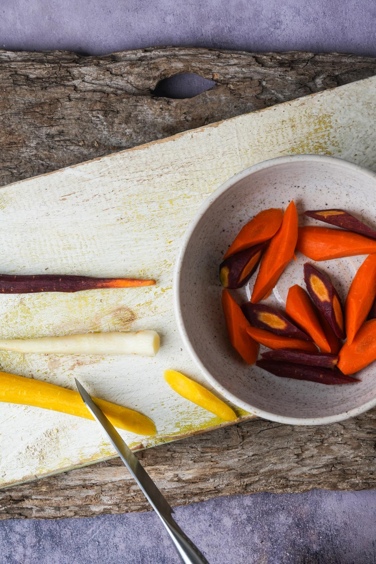 Sliced colorful carrots on a white cutting board and in a bowl, with a knife placed nearby.