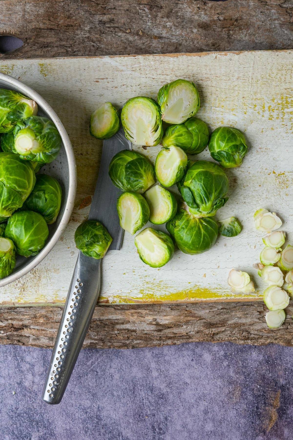 Brussels sprouts are scattered on a cutting board with a knife; some are halved, others whole, showcasing fresh green layers. A bowl with more sprouts is on the left.