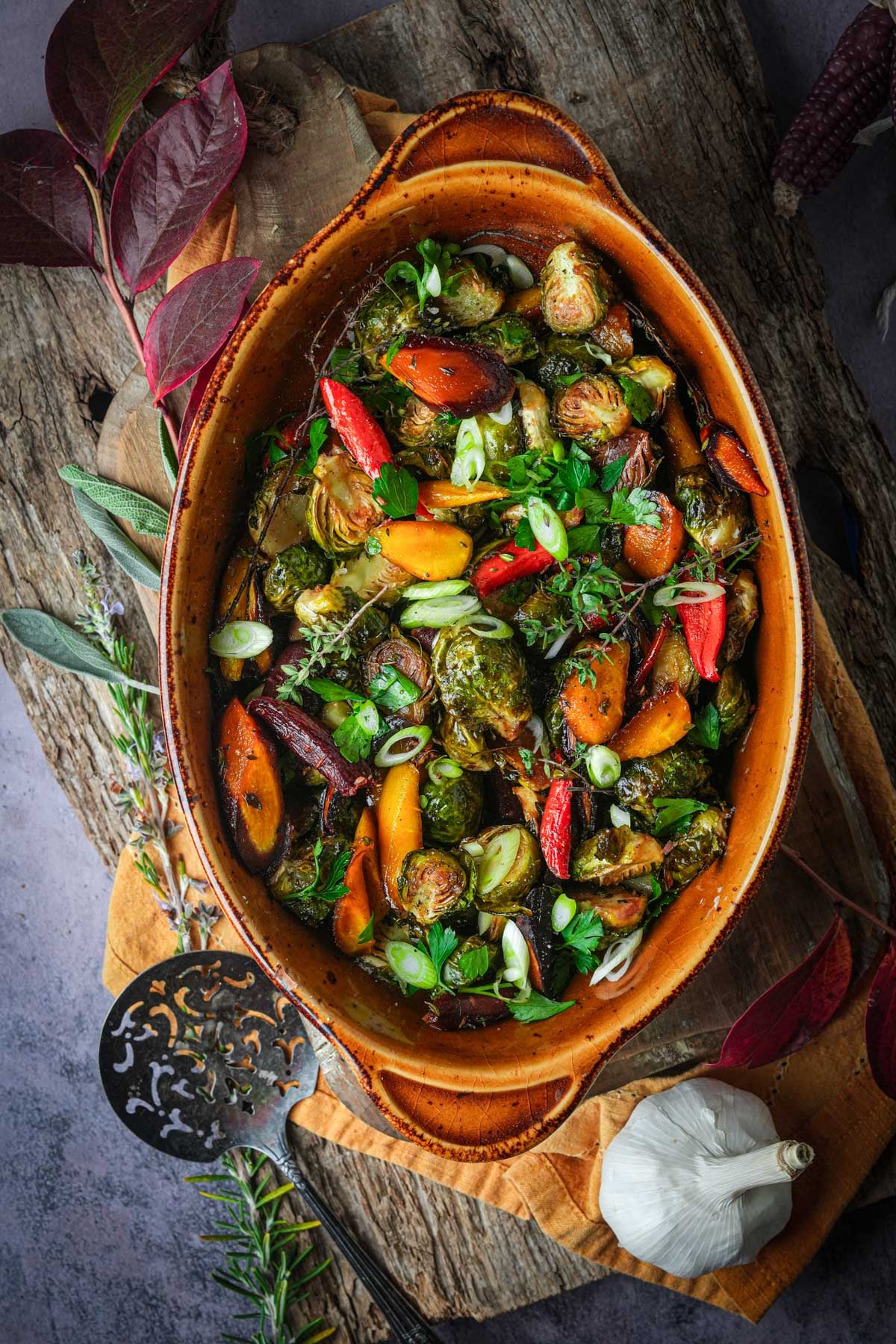 Oval dish filled with roasted Brussels sprouts and carrots, garnished with herbs, on a rustic wooden board with decorative leaves and garlic nearby.