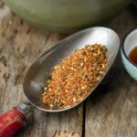 A metal spoon filled with Shichimi Togarashi placed on a wooden surface, next to a small bowl with tamari.