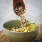 A wooden spoon sprinkles furikake seasoning over a bowl of avocado, rice, and greens.