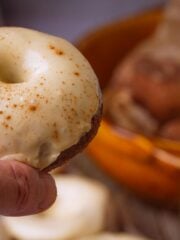 A hand holds a glazed vegan gingerbread donut with cinnamon sprinkled on top, with a wooden bowl of baked goods in the blurred background.