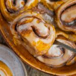 A vegan pumpkin cinnamon roll topped with maple cream cheese icing being lifted from a baking dish filled with more rolls.