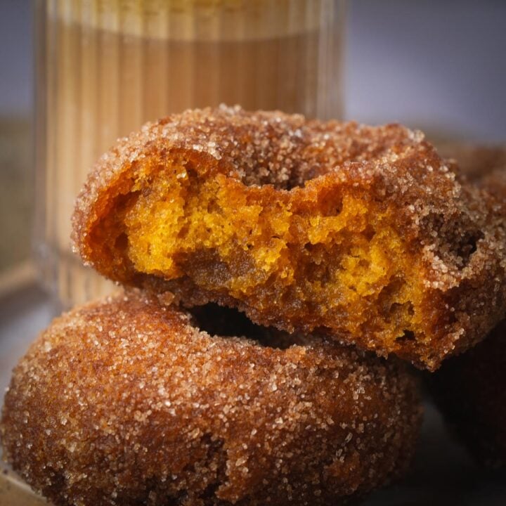 Close-up of cinnamon sugar-covered vegan pumpkin donuts stacked with a bite taken out of the top one, in front of a glass of iced coffee.