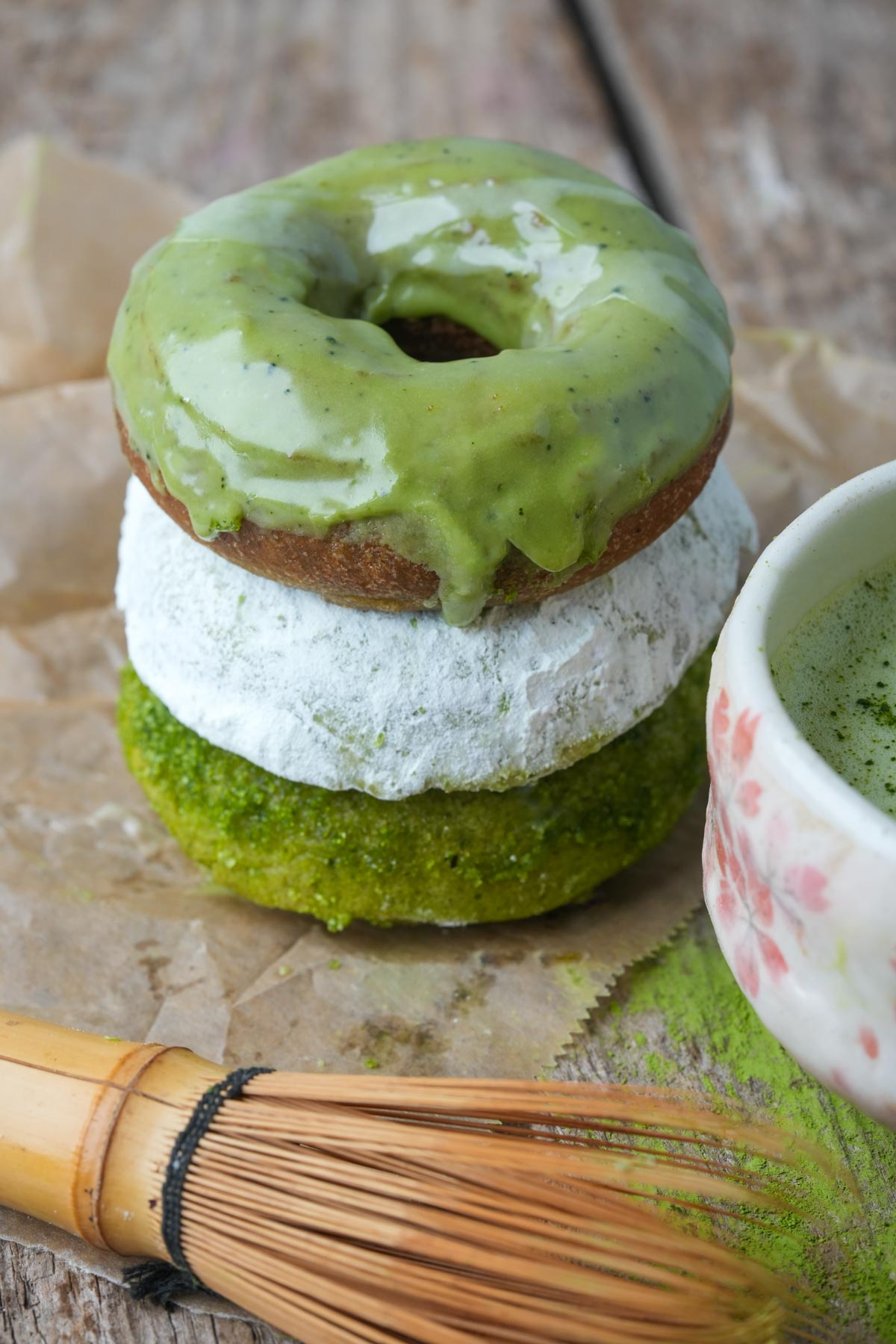A stack of three vegan matcha donuts with matcha glaze, powdered sugar, and green matcha sugar coating next to a cup and a bamboo whisk on a wooden surface.