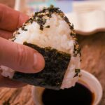 A hand holds a triangular onigiri rice ball covered with seaweed and sesame seeds over a small bowl of soy sauce.