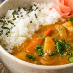A bowl of vegan Japanese curry rice with chunks of potatoes and carrots, garnished with chopped green onions, furikake, and pink pickled ginger on the side.
