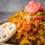 A dish of stir-fried vegan yakisoba noodles with sesame seeds, sliced lotus root, green onions, and pickled ginger.