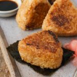 Grilled yaki onigiri on a plate with nori and pickled ginger, accompanied by a small dish of soy sauce.