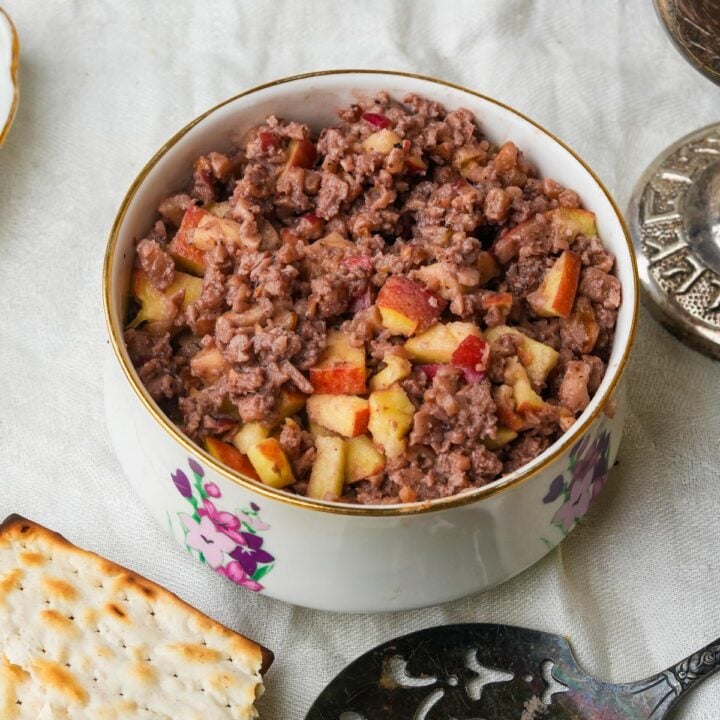 A bowl filled with charoset made from apples and minced nuts, resting on a white cloth. A cracker and a silver utensil are nearby.