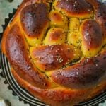A close-up of a glazed, braided round challah bread topped with sesame seeds, placed on a patterned tray.