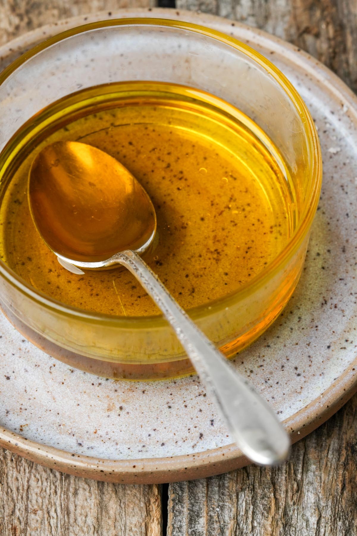 A glass bowl of golden glaze with a metal spoon inside, placed on a rustic wooden surface.