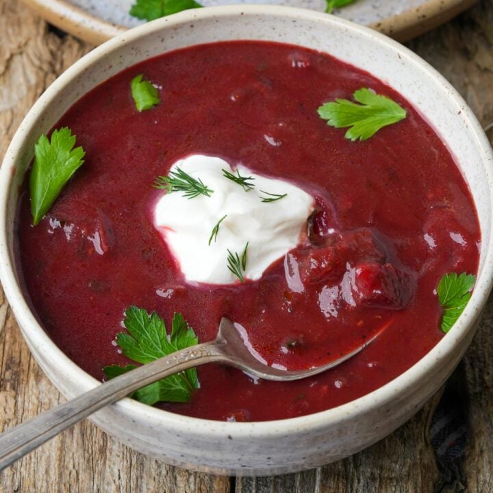 A bowl of red vegan borscht soup with a dollop of tofu sour cream and dill on top. Garnished with parsley leaves, and a spoon rests inside the bowl.