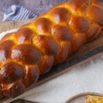 A golden-brown braided loaf of vegan challah bread on a wooden board with a blue and white cloth partially visible.