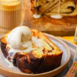 A slice of vegan cinnamon babka with a scoop of vanilla ice cream on a plate, with a glass of iced coffee and more bread in the background.