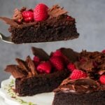 A slice of vegan flourless chocolate cake topped with raspberries and chocolate shavings is being lifted from a cake stand with a spatula.